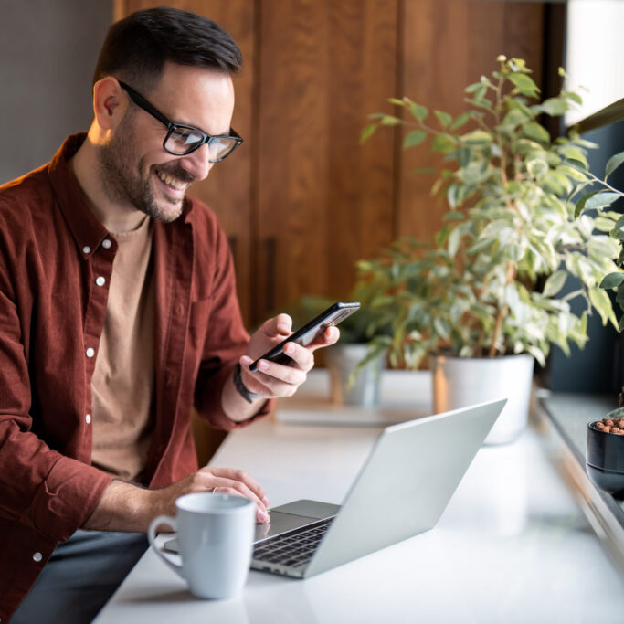 Satisfied modern millennial man in stylish casual clothes using smartphone and laptop computer for electronic banking, making reservation, online shopping and payments while spending time at home.