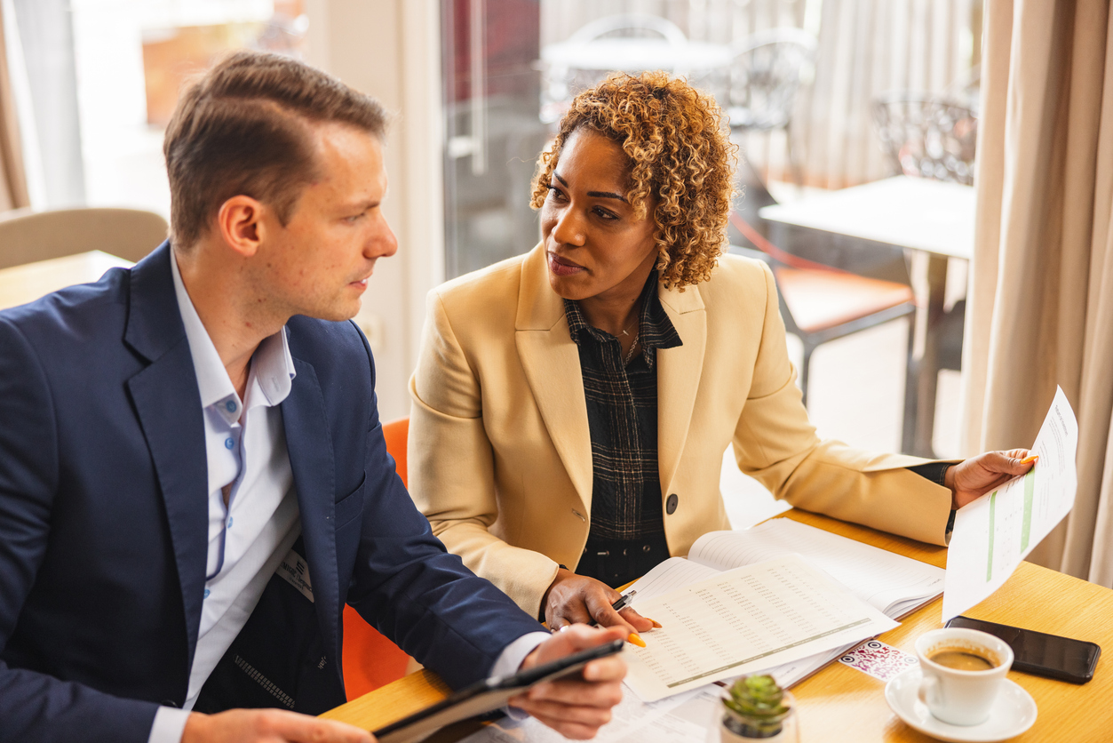 A professional mid-adult businessman looking at the documents together with his female colleague. They are sitting in a corporate cafeteria and discussing the numbers and strategies. They look serious and focused. They are wearing businesswear.